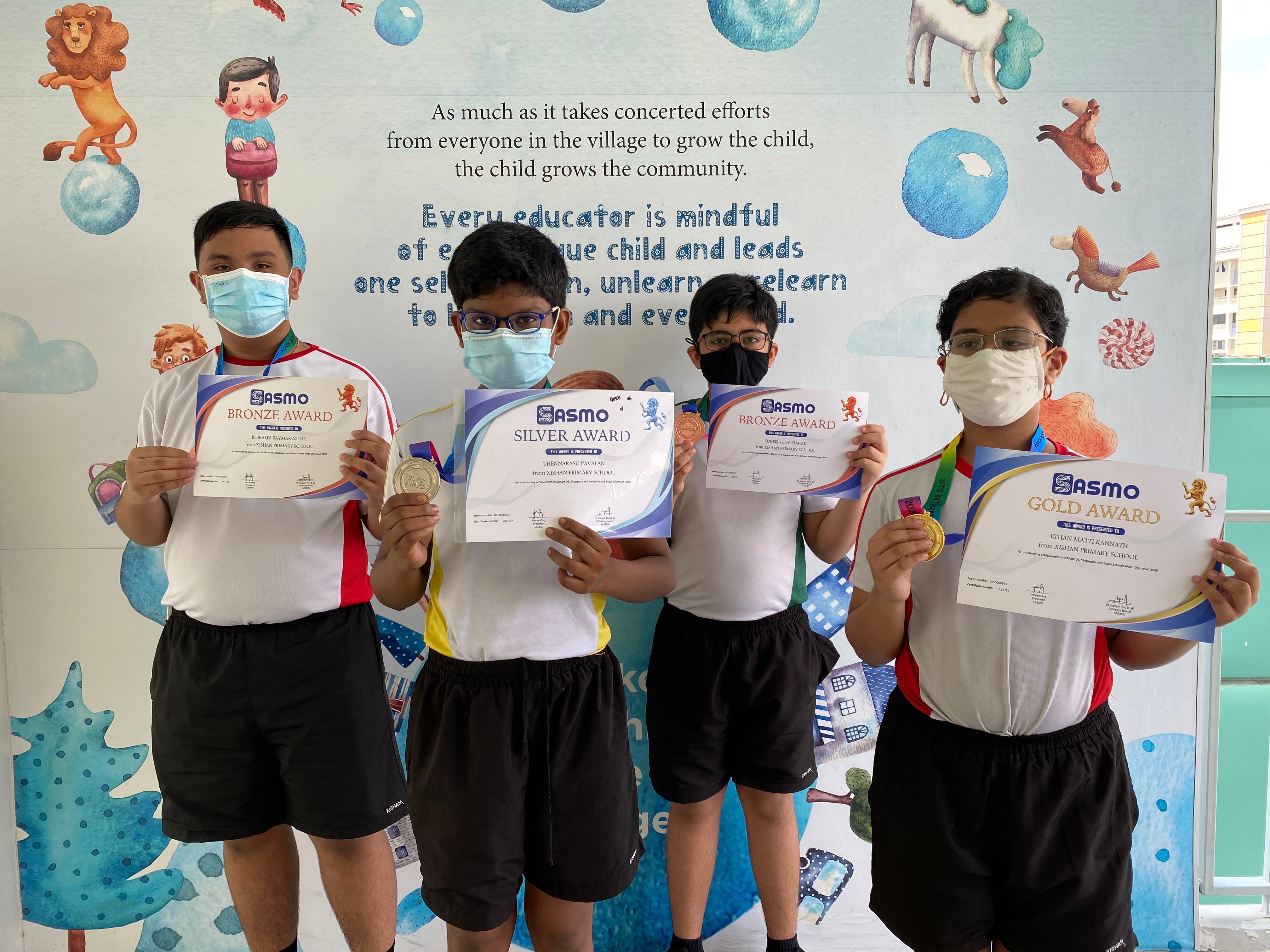Four masked students hold SASMO award certificates: two Bronze, one Silver, and one Gold, in front of a colorful mural.